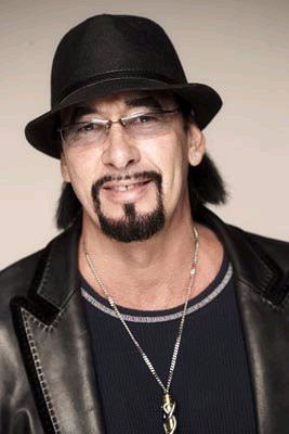 Dark haired man with a black hat, glasses and a goatee, wearing a black shirt with a black leather jacket
